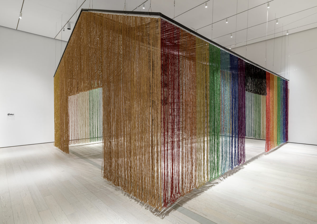 Long strands of various colors form a structure that hangs in an all-white room.
